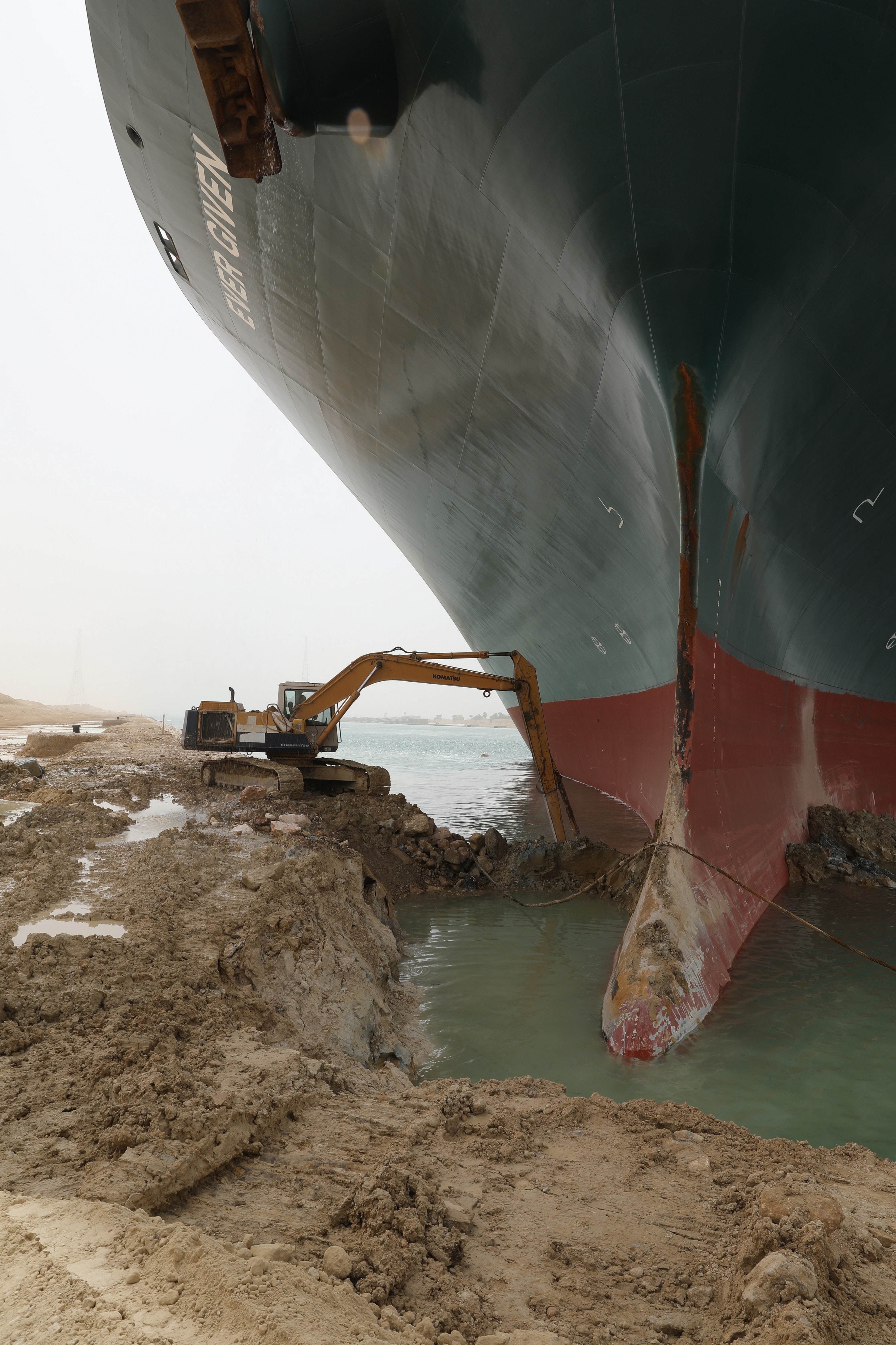 A digger extracting soil at the base of a large ship's hull